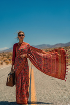 Woman in red patterned saree and sunglasses standing on a road in a desert landscape