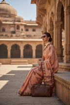 Woman in peach saree with geometric patterns, sitting in a traditional courtyard with a leather handbag