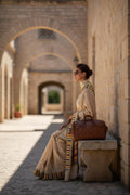 Woman in elegant cream saree with striped border, sunglasses, and brown leather bag sitting on stone bench outdoors