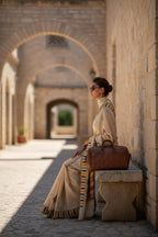 Woman in elegant cream saree with striped border, sunglasses, and brown leather bag sitting on stone bench outdoors