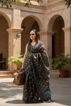 Woman in a black and gold saree with sunglasses, holding a brown handbag outdoors